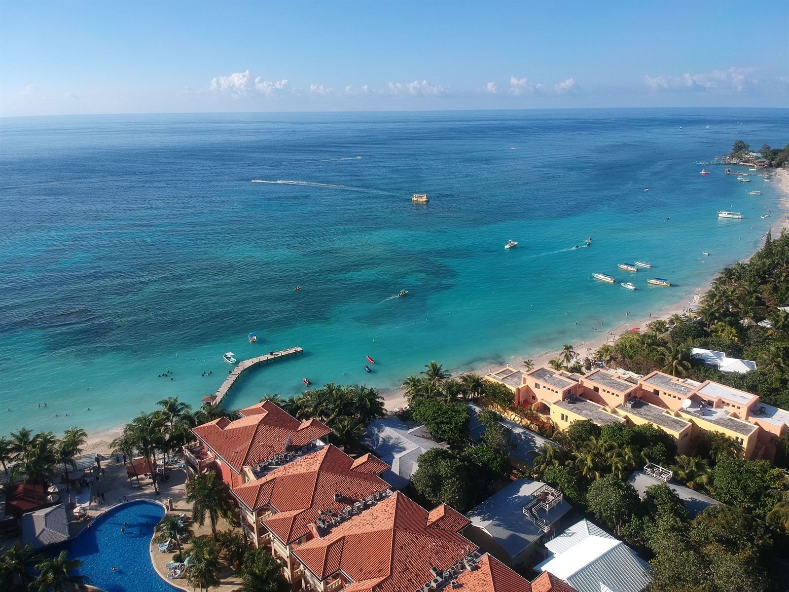 Aerial view of red-roofed resort buildings, a pool and a small pier beside turquoise water on Roatan